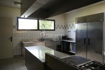 Cuisine professionnelle avec plan de travail en inox, cuisinière, évier, hotte et équipements muraux dans un espace carrelé.