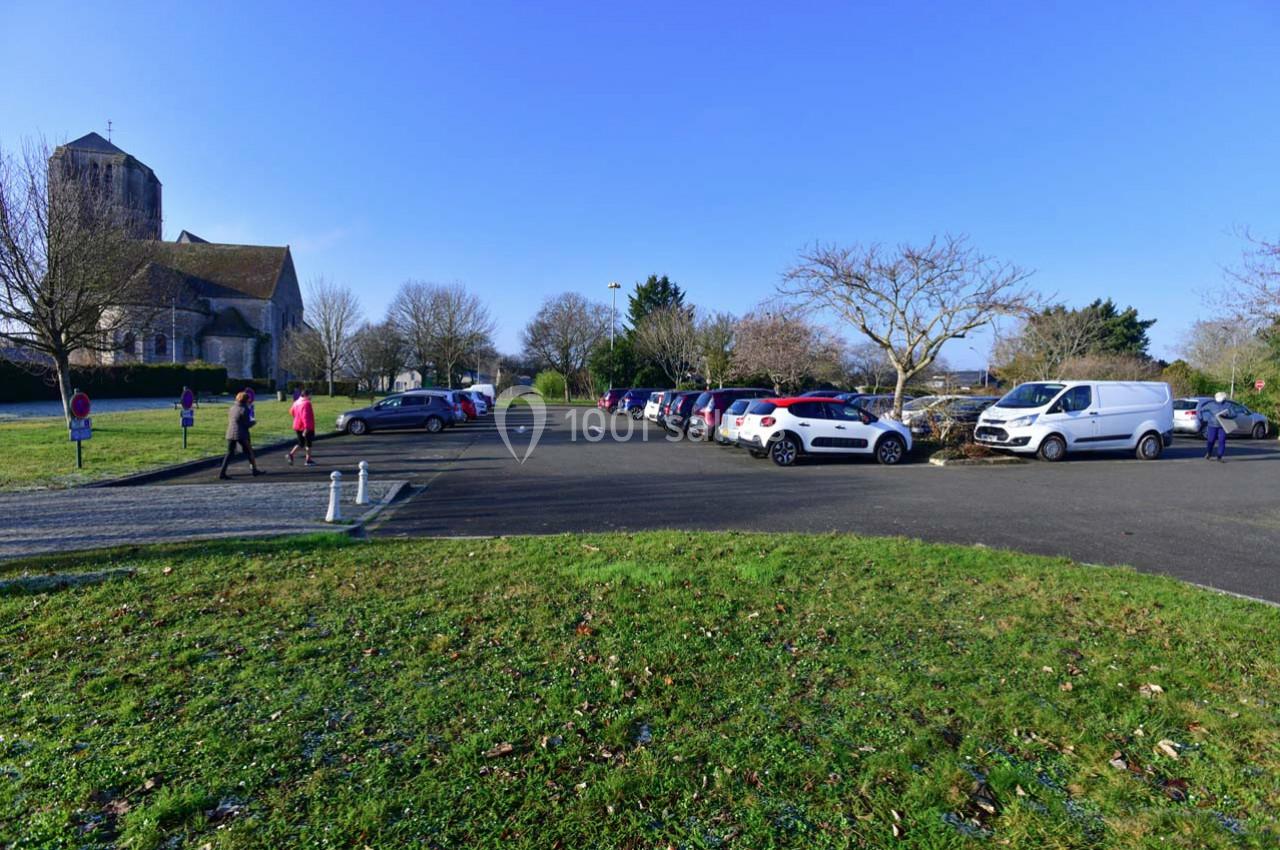 Parking extérieur avec plusieurs voitures, des piétons et un bâtiment ancien en pierre à l'arrière-plan.