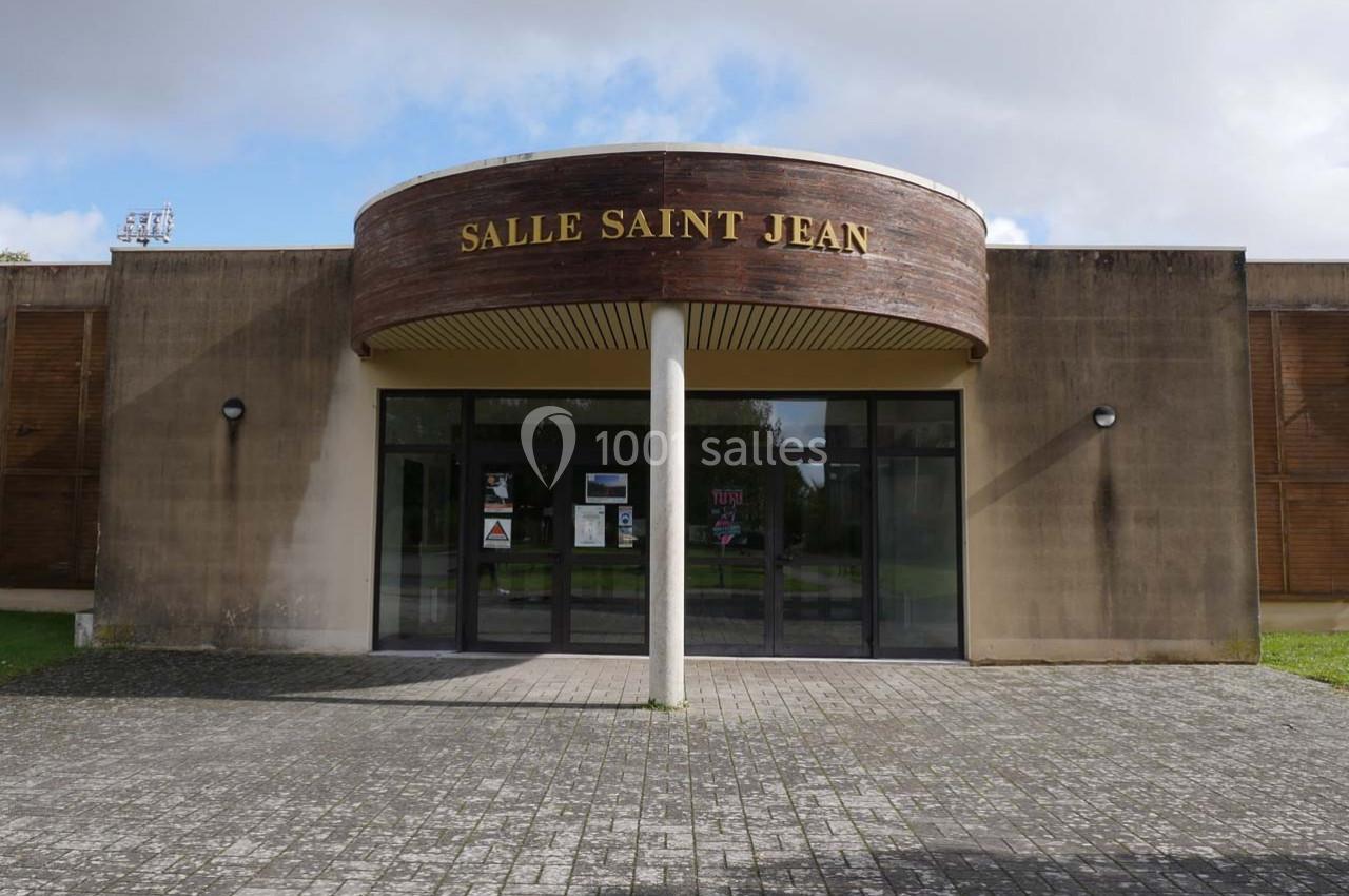 Façade d'un bâtiment avec une entrée vitrée, un panneau ’Salle Saint Jean’ et un sol pavé devant.