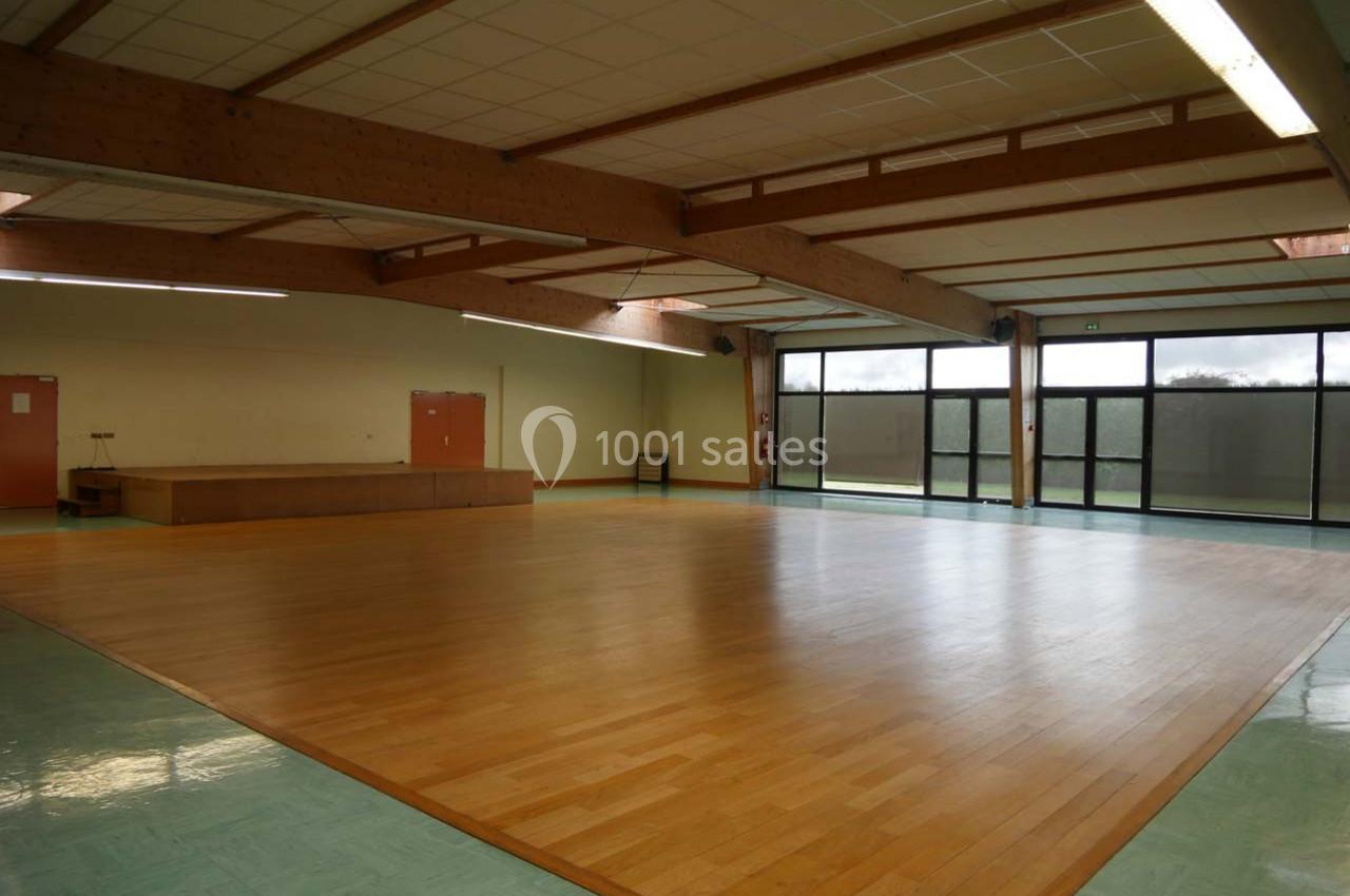 Salle polyvalente vide avec parquet en bois, murs clairs, plafond à poutres apparentes et grandes baies vitrées.