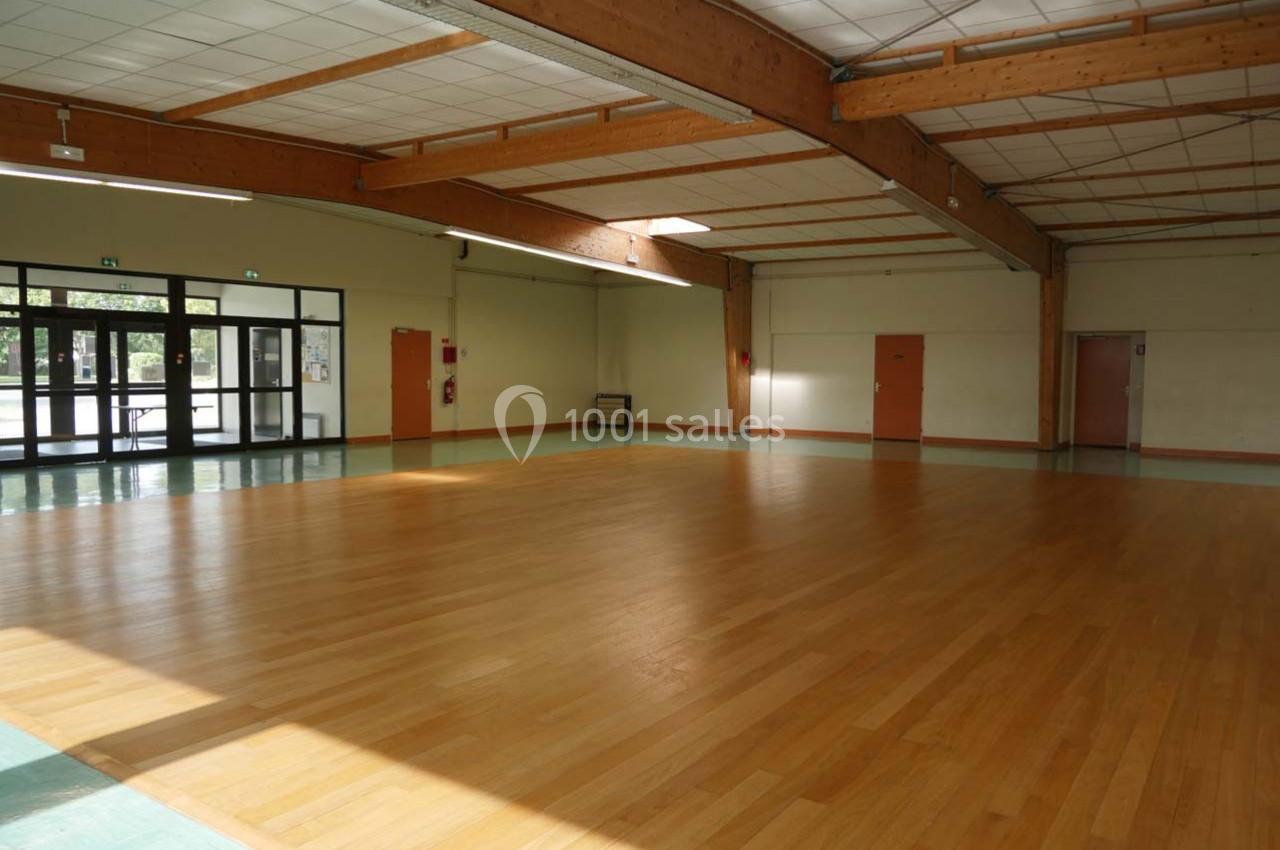 Salle vide avec parquet en bois, murs clairs, plafond avec poutres apparentes et grandes baies vitrées.