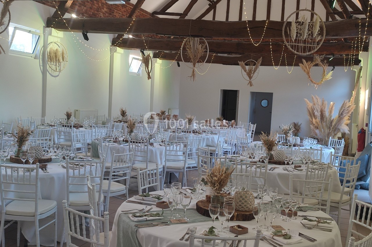 Salle de réception décorée avec des tables rondes, nappes blanches, chaises blanches et ornements végétaux suspendus.