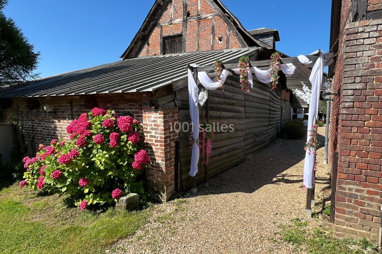 Allée en gravier bordée d'un bâtiment en bois et briques, décorée de fleurs et de voilages blancs.