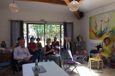 Des personnes assises sur des tapis et des chaises dans une salle lumineuse avec un tableau et des rideaux.