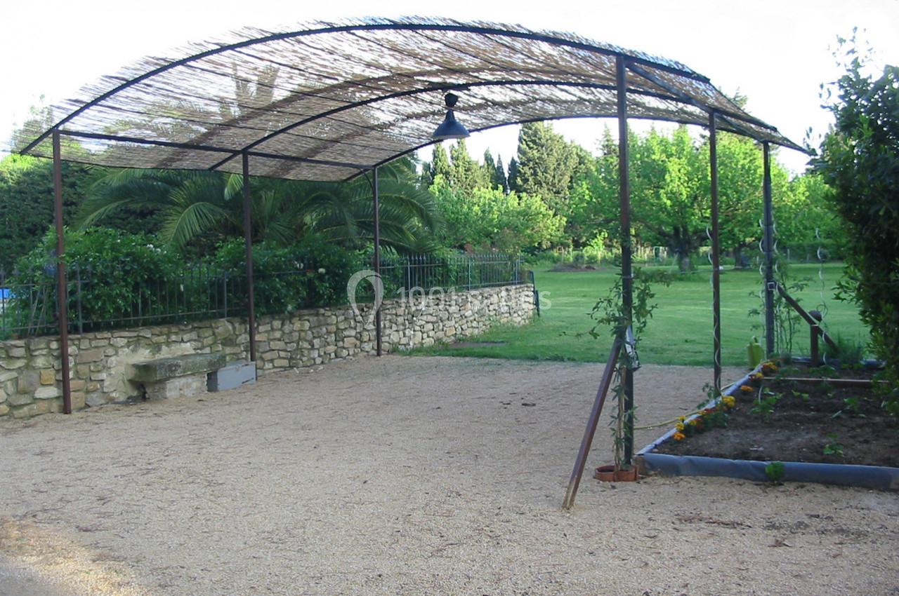 Abri en structure métallique avec toit en canisse, situé sur une allée gravillonnée près d'un jardin verdoyant.
