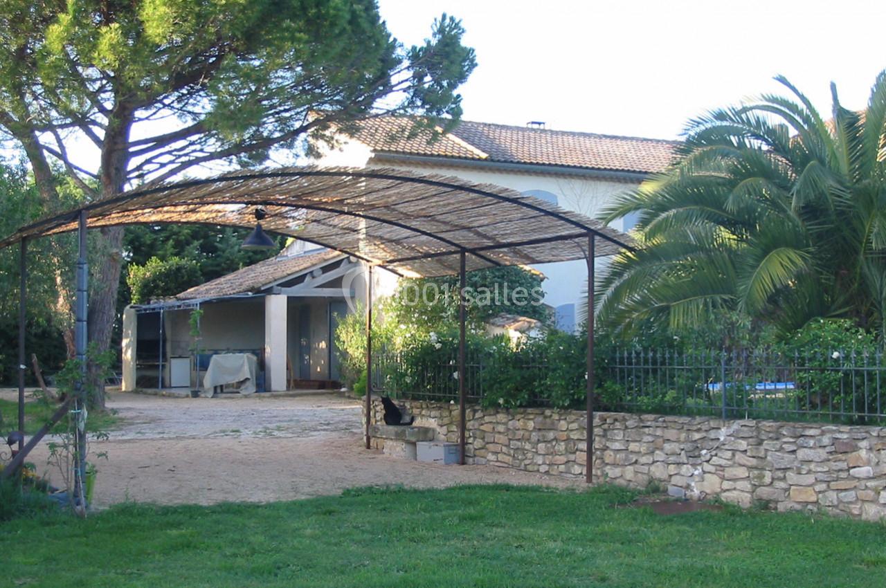 Abri métallique devant une maison entourée de végétation, avec un arbre, un palmier et une clôture en pierre.