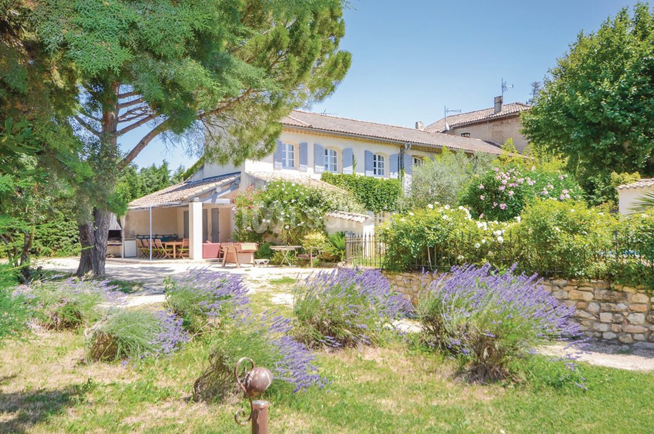Maison provençale entourée de lavandes, arbres et jardin fleuri sous un ciel bleu clair.