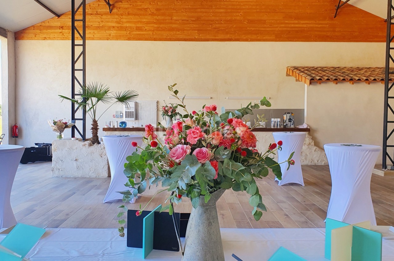 Salle lumineuse avec des tables hautes drapées de blanc et un grand bouquet de fleurs roses au premier plan.