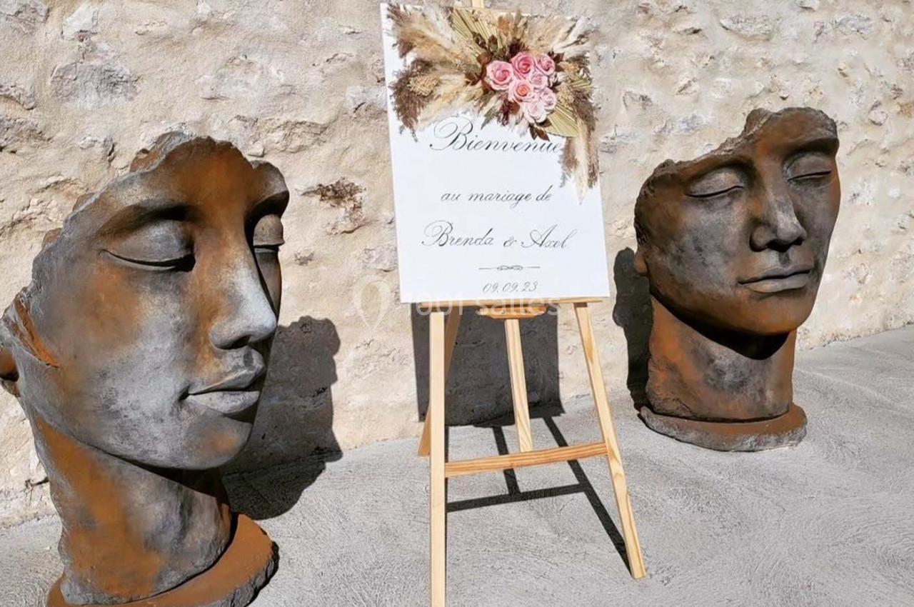 Deux sculptures de visages en pierre encadrent un chevalet avec une affiche de mariage décorée de fleurs séchées.
