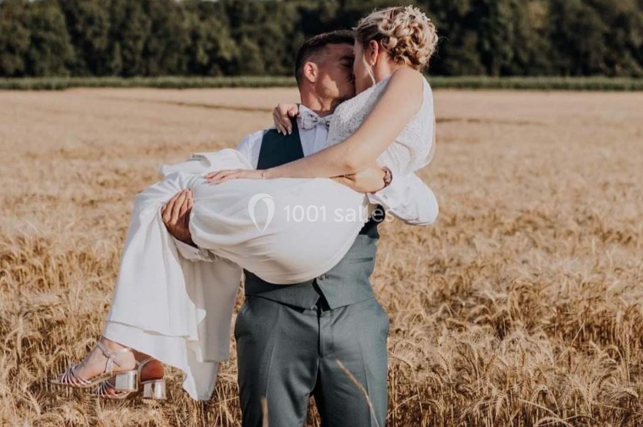 Un homme en costume porte une femme en robe blanche dans un champ de blé sous un ciel dégagé.