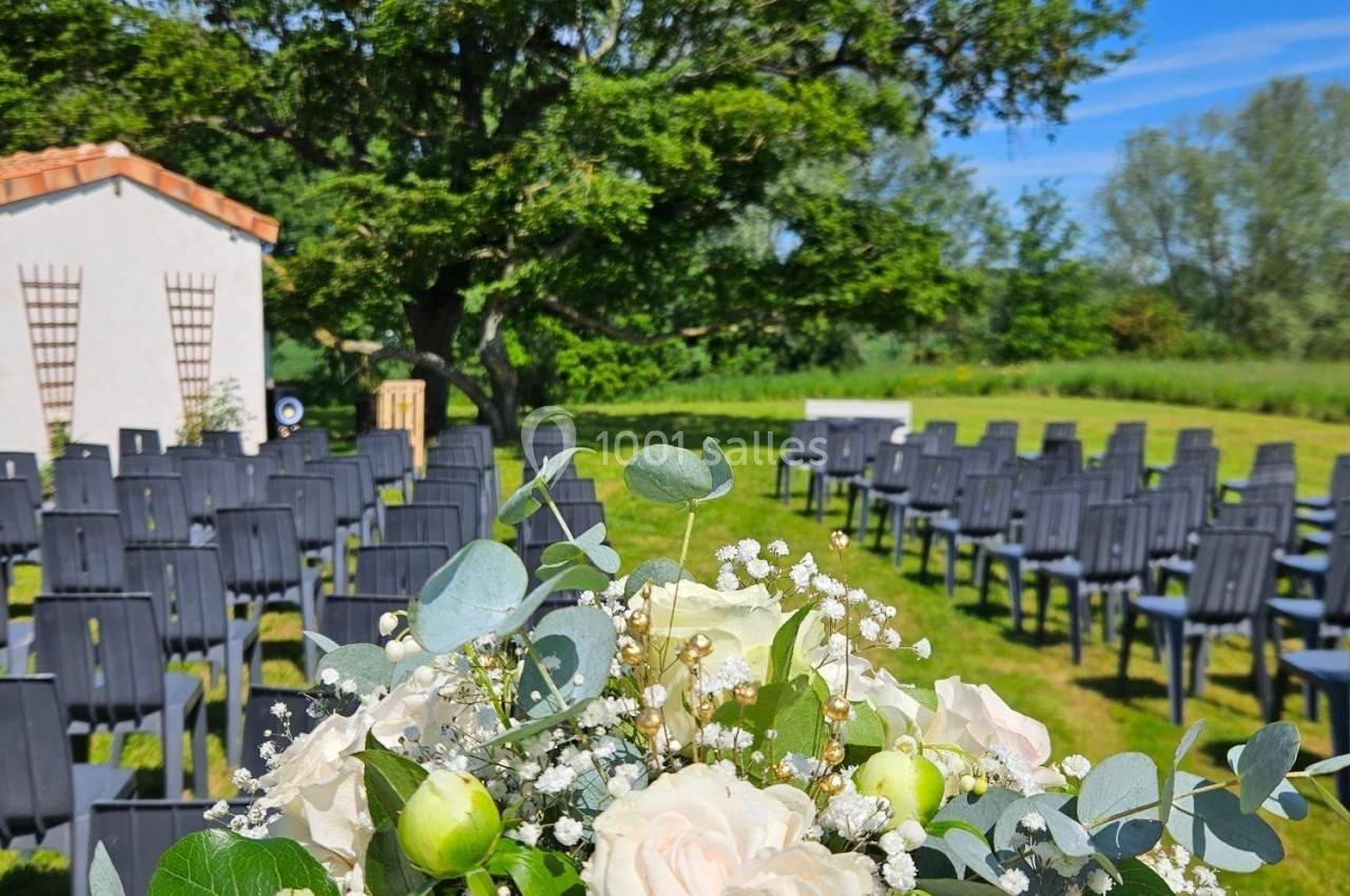 Bouquet de fleurs blanches et vertes au premier plan, avec des chaises disposées en extérieur sous un grand arbre.
