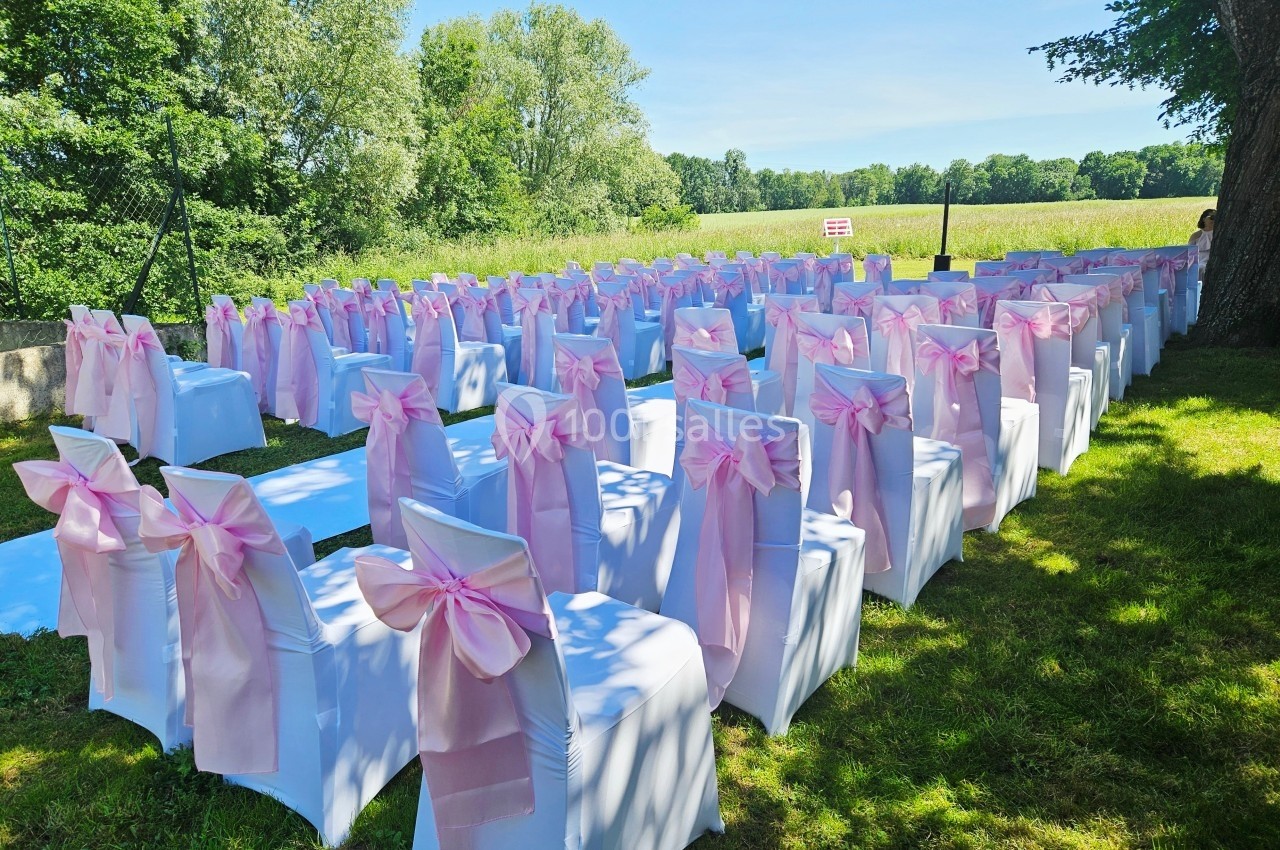 Chaises blanches décorées de nœuds roses alignées en extérieur pour une cérémonie sur une pelouse ensoleillée.