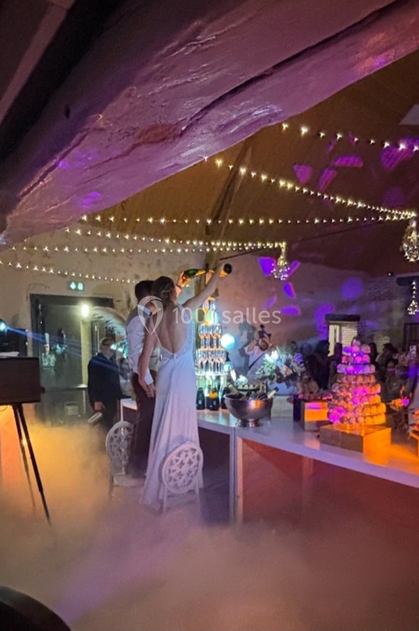 Un couple célèbre son mariage en sabrant une bouteille de champagne dans une salle décorée de guirlandes lumineuses.