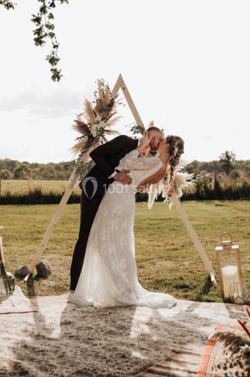 Un couple en tenue de mariage s'embrasse devant une arche décorée, dans un cadre champêtre en plein air.
