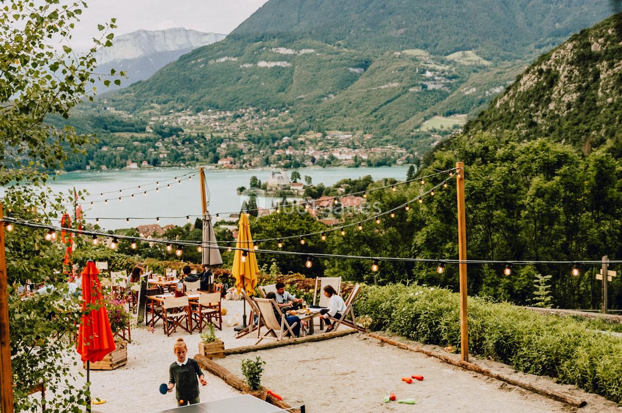 Enfants jouant au ping-pong sur une terrasse avec vue sur un lac, des montagnes et des tables de café en arrière-plan.