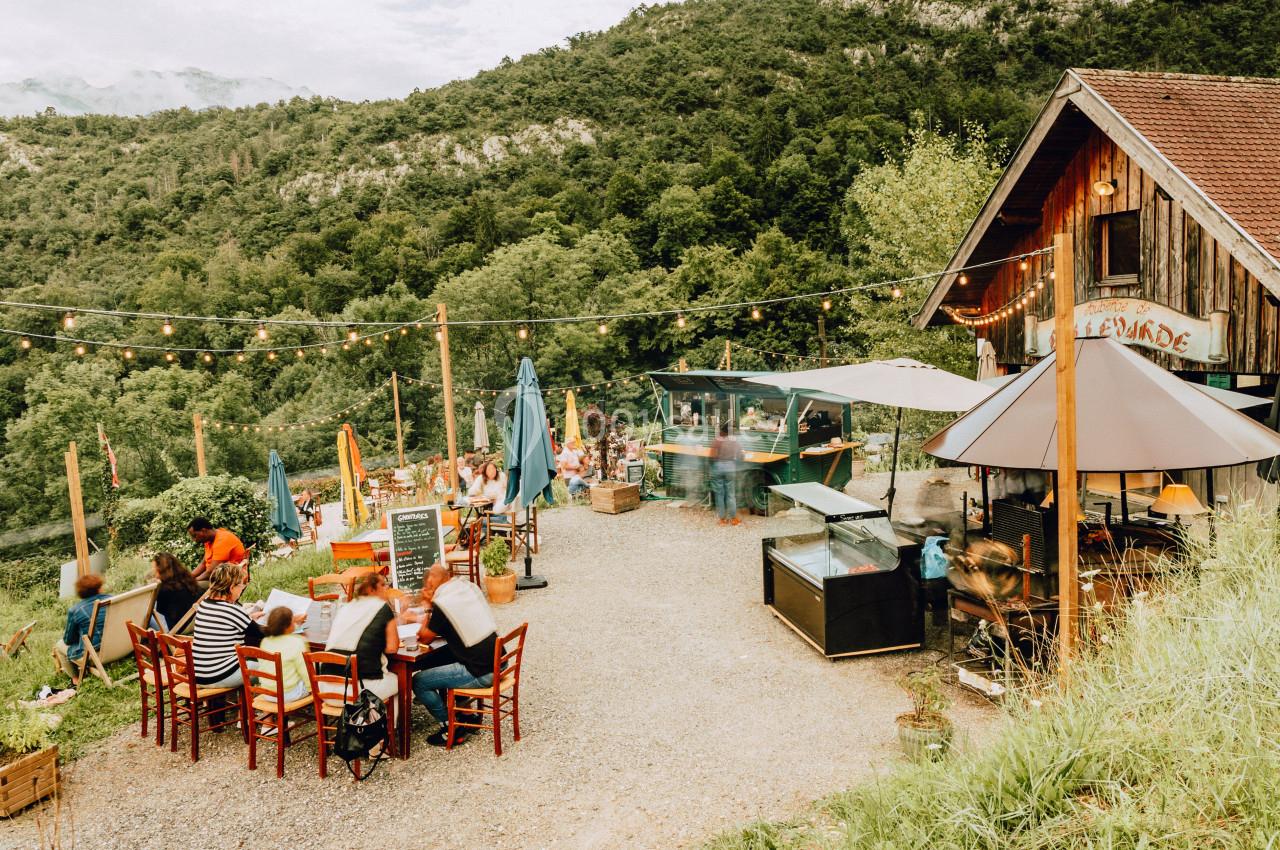 Terrasse extérieure d'un restaurant rural avec des clients assis, entourée de verdure et de collines.