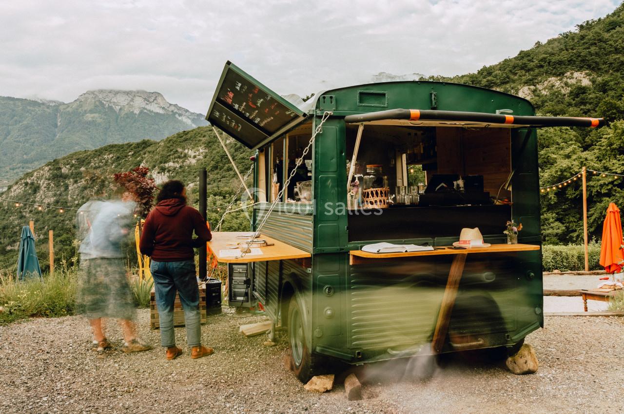 Camion-restaurant vert aménagé en plein air, entouré de montagnes et de clients en attente.