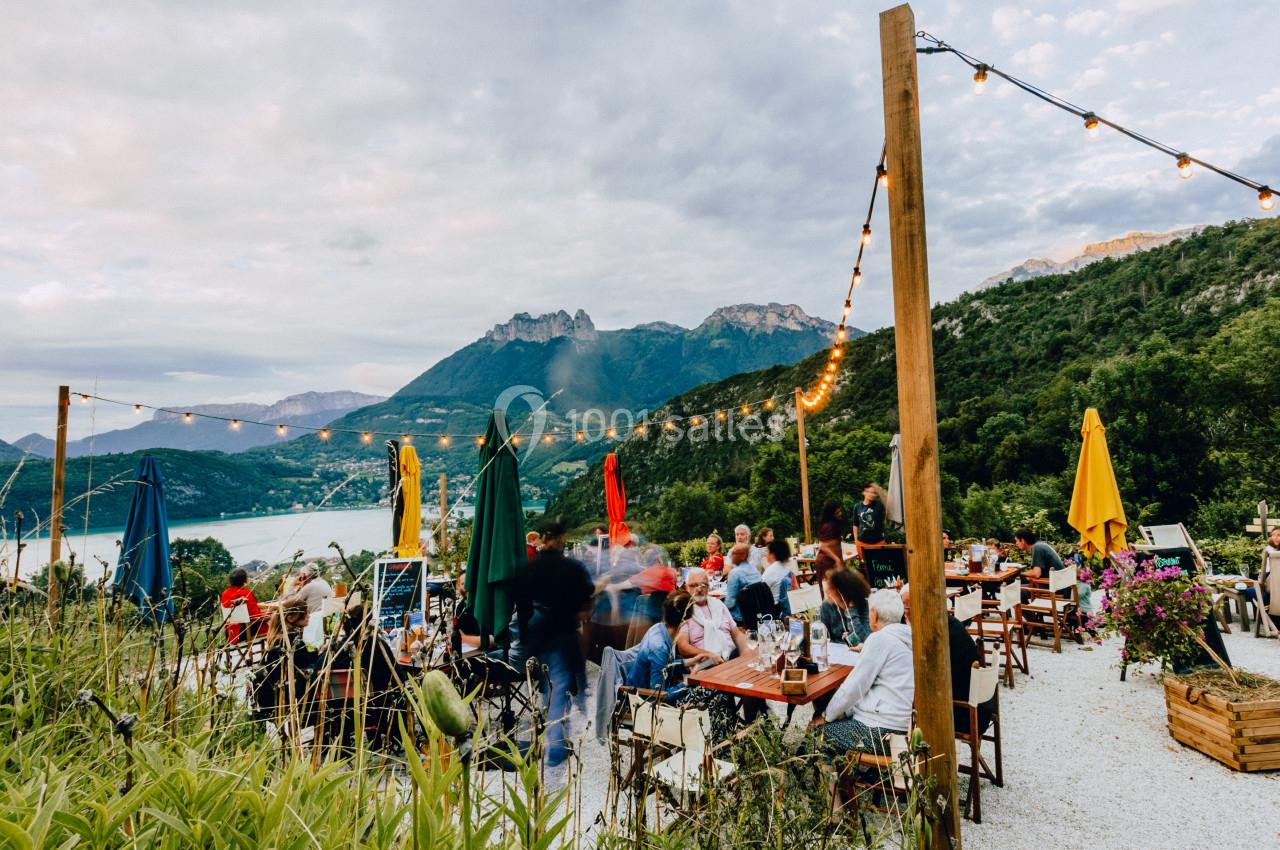 Terrasse animée avec des tables et des parasols colorés, vue sur un lac entouré de montagnes et de végétation.