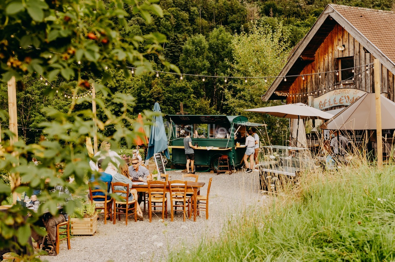 Personnes assises et debout dans un café en plein air, entouré de verdure et de bâtiments en bois.