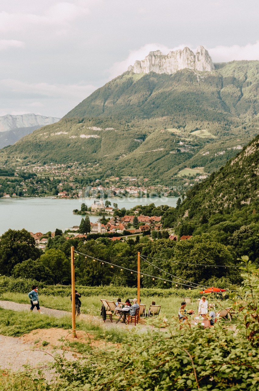 Vue sur un lac entouré de montagnes, avec des tables en plein air et des personnes profitant du paysage.