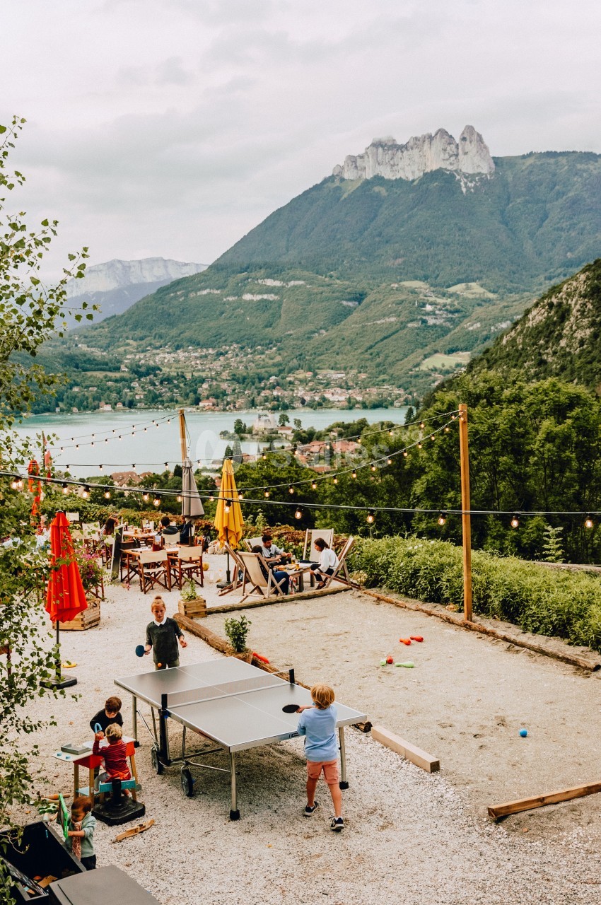 Enfants jouant au ping-pong sur une terrasse avec vue sur un lac, des montagnes et des tables de café en arrière-plan.