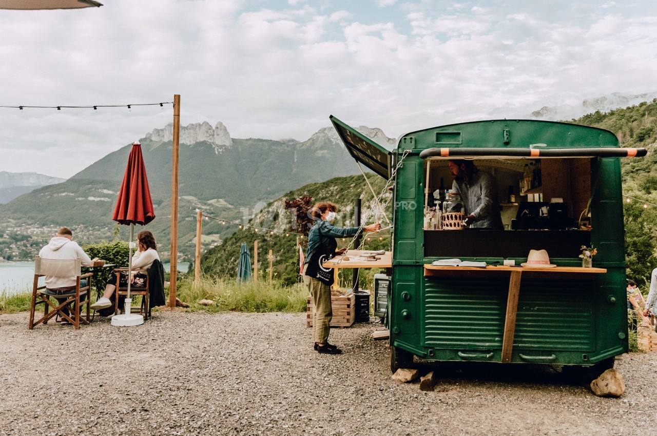 Camion-restaurant vert en plein air avec des clients assis et un paysage de montagnes en arrière-plan.