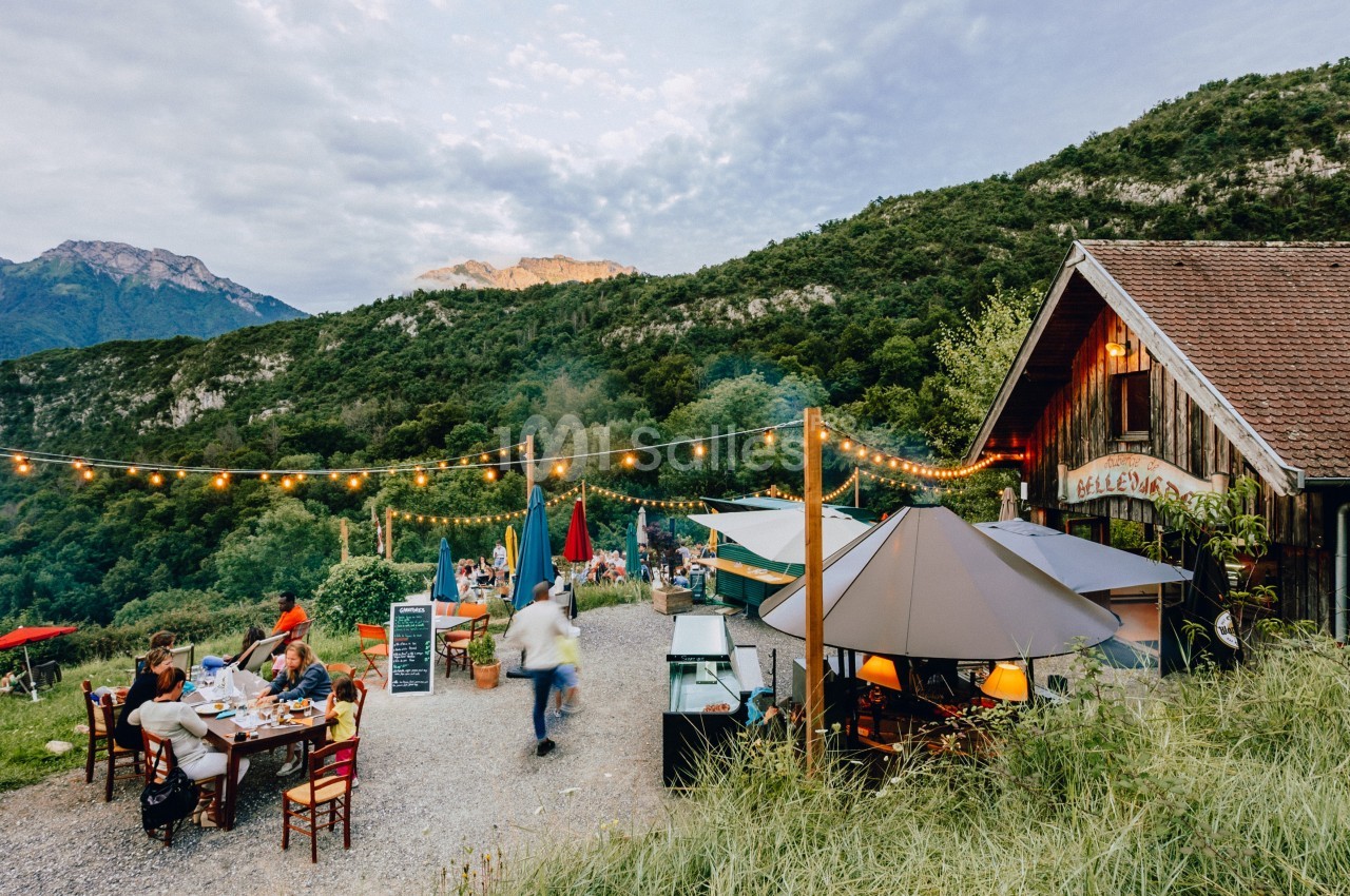 Terrasse extérieure d'un restaurant rural avec des tables, des parasols et des guirlandes lumineuses, entourée de montagnes.