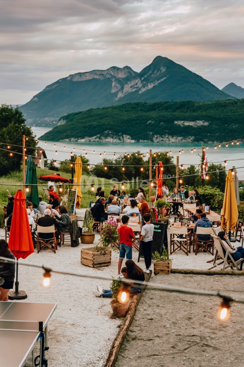 Terrasse animée en plein air avec des tables, des personnes et des lumières suspendues, vue sur un lac et des montagnes.