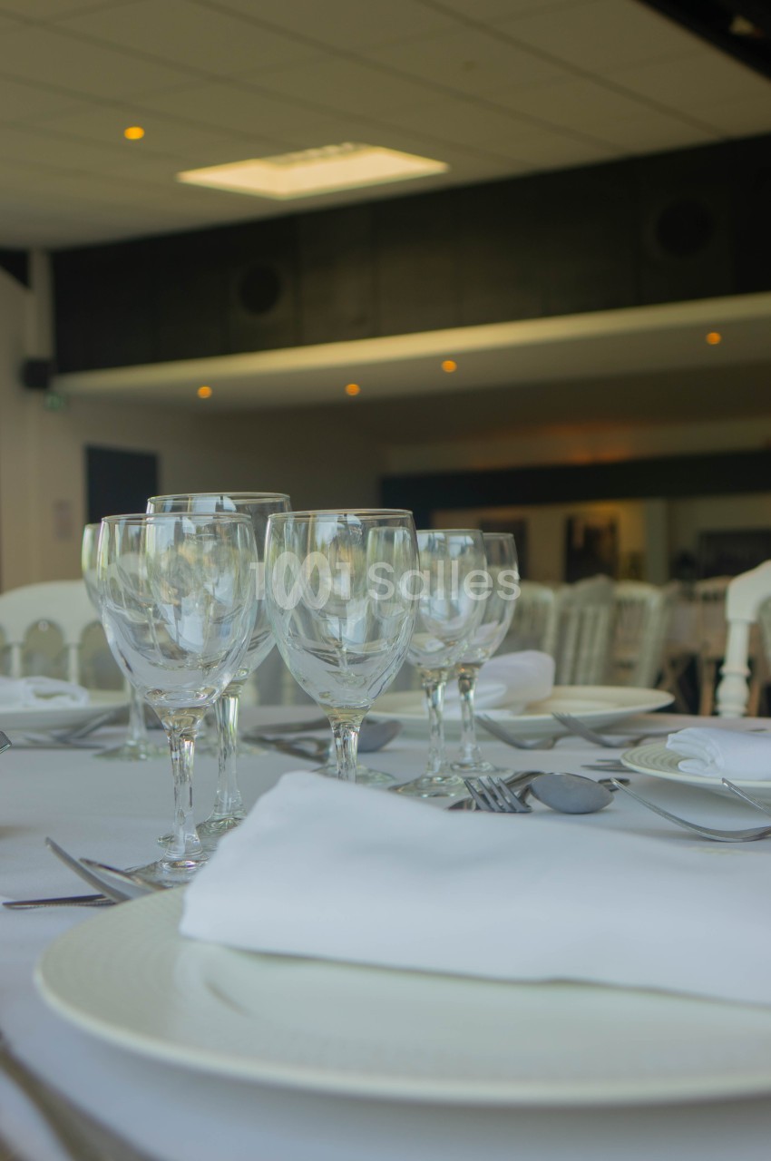 Table dressée avec des verres à pied, assiettes blanches et serviettes, dans une salle lumineuse au décor sobre.