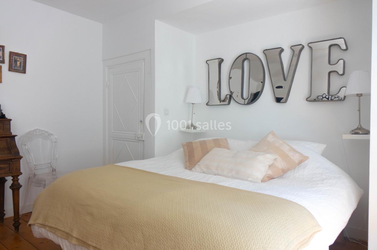 Lit double avec couverture beige, coussins assortis, mur blanc décoré de lettres métalliques formant ’LOVE’.