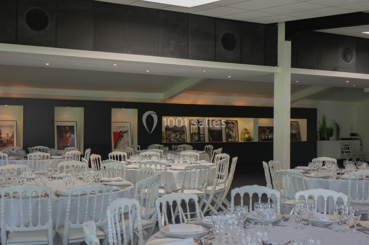 Salle de réception avec tables dressées, chaises blanches et décorations murales comprenant des photographies encadrées.