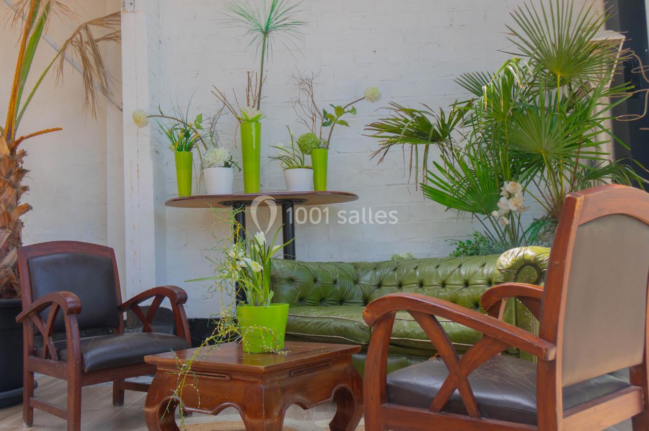 Salon lumineux avec canapé vert, fauteuils en bois, table basse et plantes vertes décoratives contre un mur blanc.