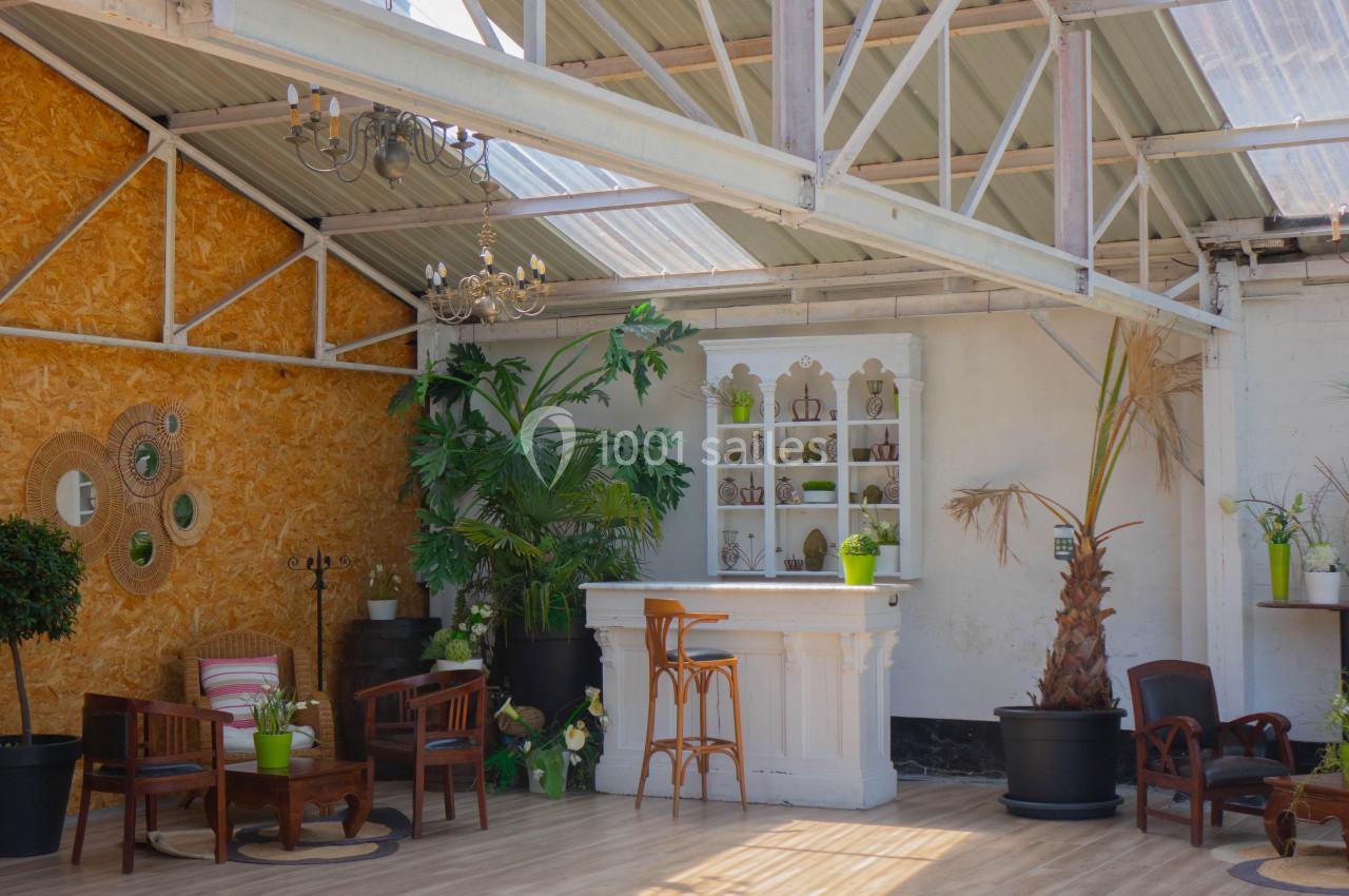 Espace lumineux avec bar blanc, plantes vertes, mobilier en bois et murs aux textures variées sous une verrière.