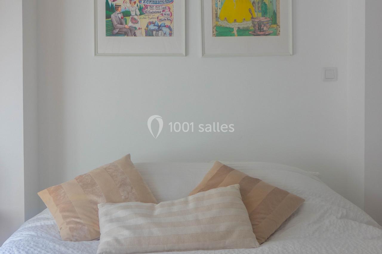 Lit double avec coussins beiges et blancs, surmonté de deux affiches encadrées accrochées à un mur blanc.