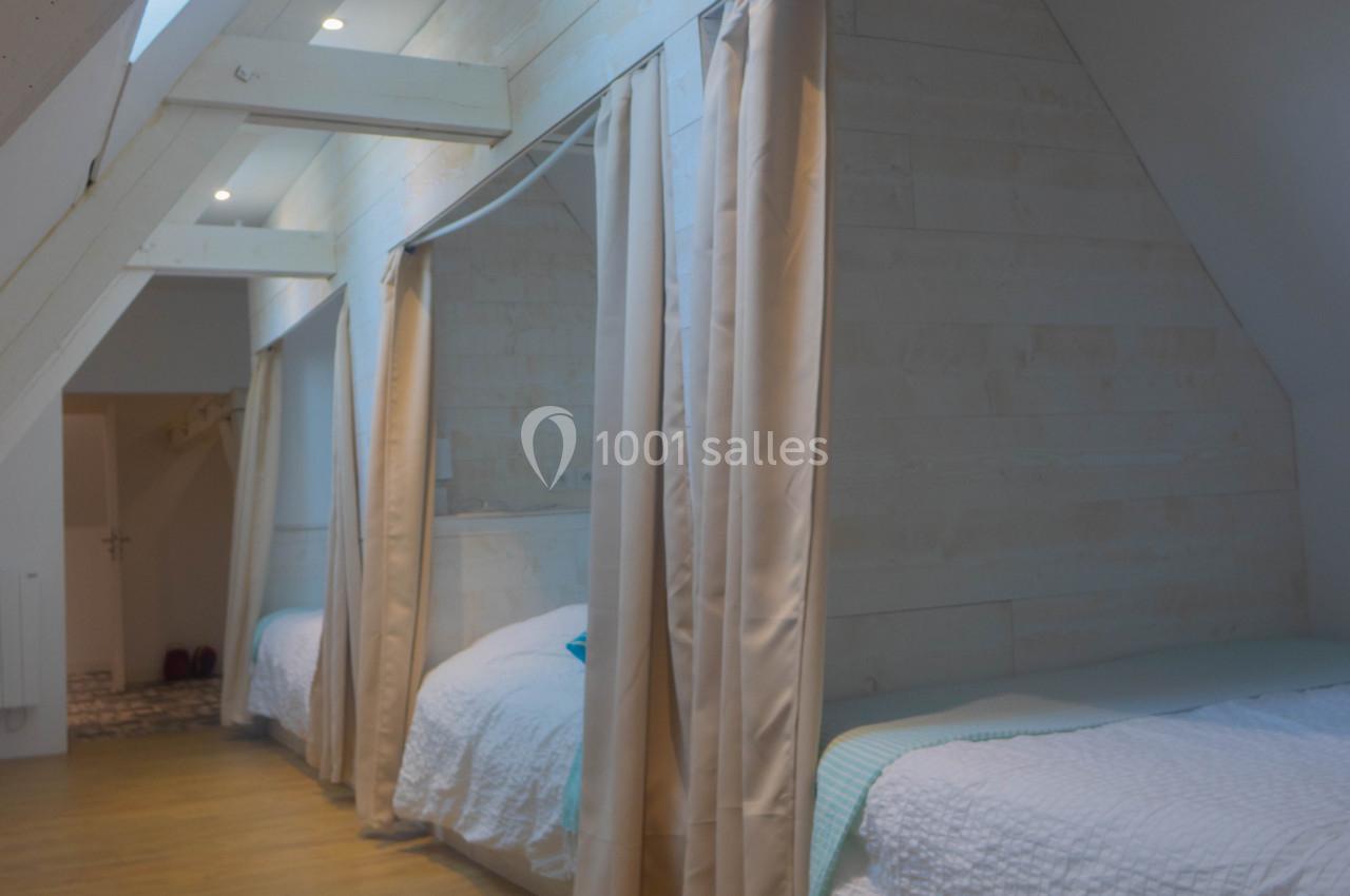 Chambre mansardée avec lits simples alignés, séparés par des rideaux, et éclairée par des fenêtres de toit.