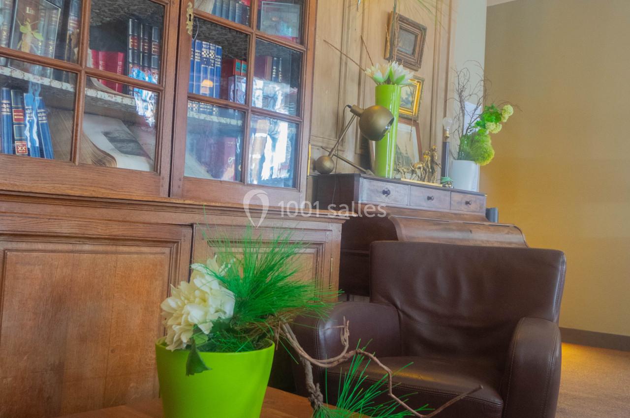Salon avec fauteuil en cuir, table en bois, plante verte en pot et bibliothèque vitrée contenant des livres.