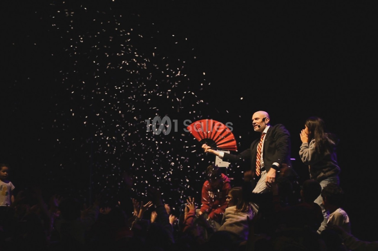 Un homme en costume avec un éventail rouge sur scène, entouré d'enfants et de confettis tombant dans l'obscurité.
