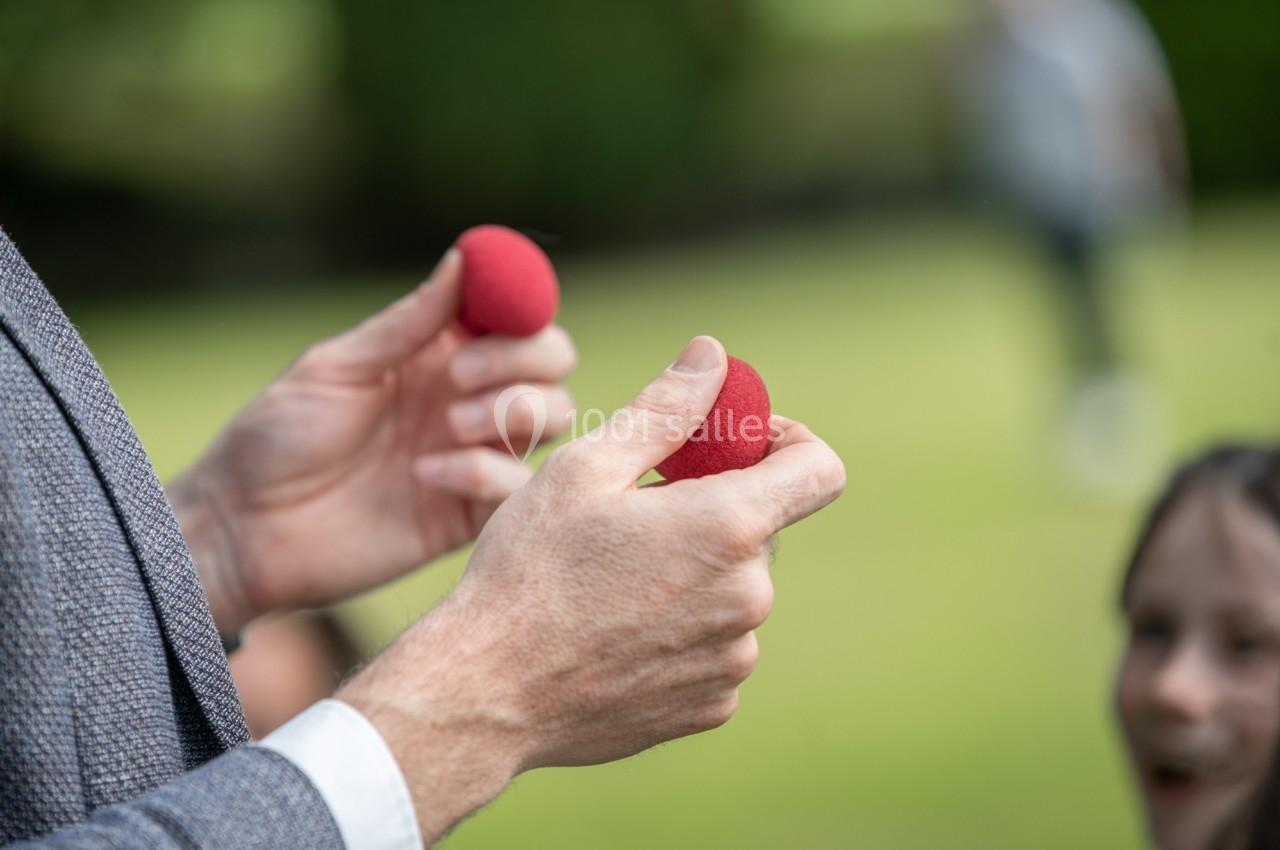 Deux mains tenant des balles rouges en mousse dans un jardin, avec des personnes floues en arrière-plan.