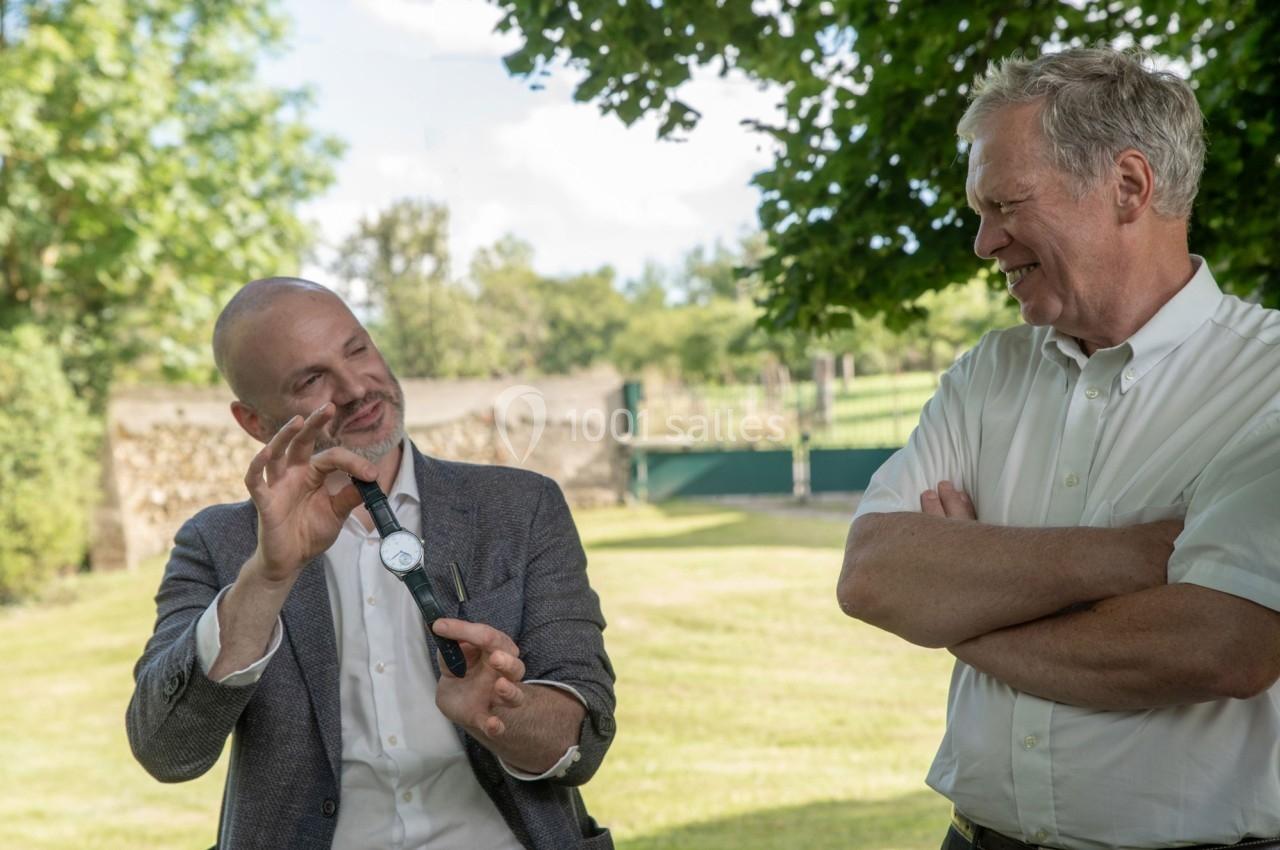Deux hommes discutent dehors dans un jardin, l'un tenant une montre et l'autre écoutant en souriant.