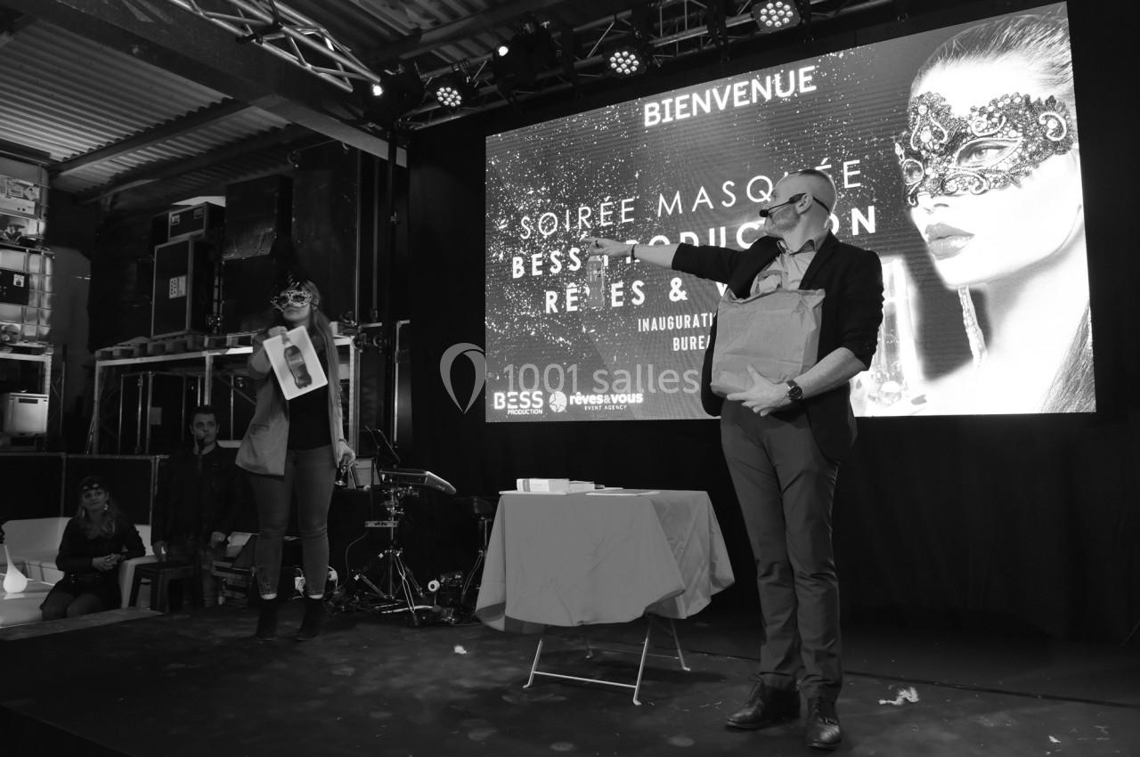 Deux personnes sur scène lors d'une soirée masquée, avec un écran affichant ’Bienvenue’ en arrière-plan.