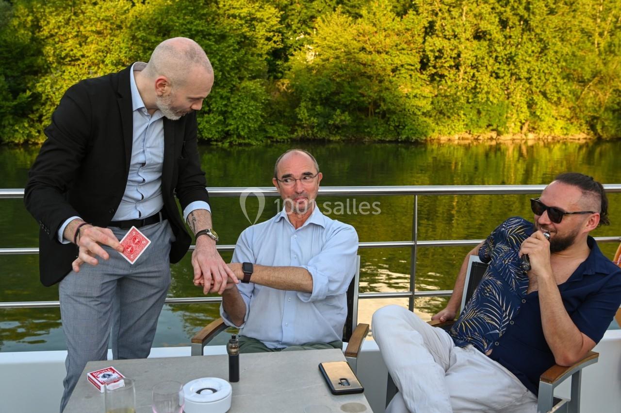 Un magicien réalise un tour de cartes pour deux hommes assis sur une terrasse au bord d'une rivière.