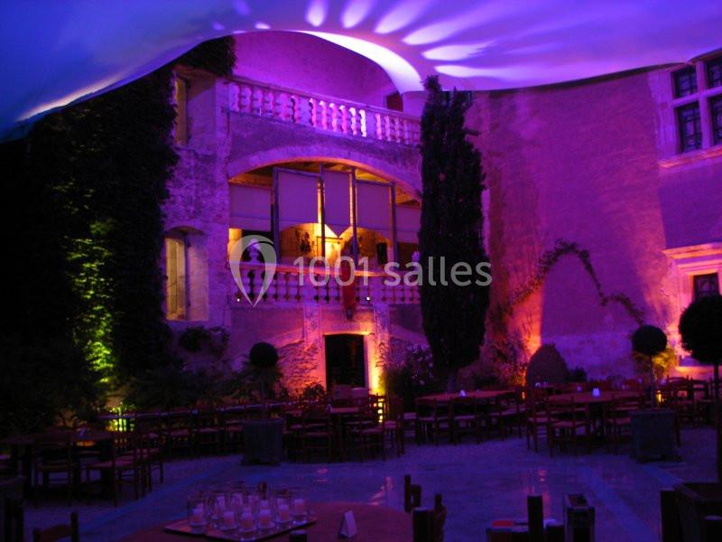 Cour intérieure illuminée en violet avec des tables dressées et des murs en pierre ornés de végétation.