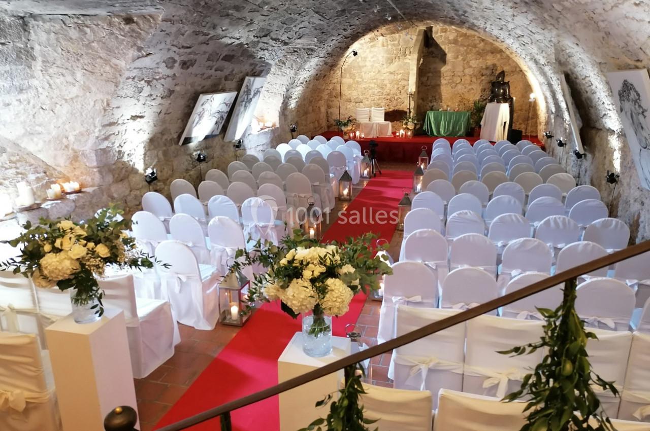 Salle voûtée en pierre aménagée pour une cérémonie, avec des chaises blanches alignées et une allée centrale rouge.