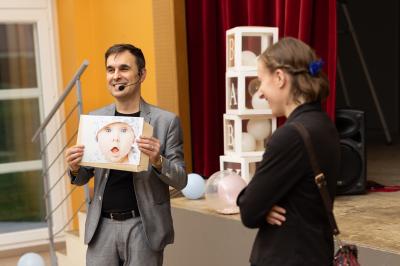 Un homme tenant une photo d'un bébé s'adresse à une femme dans une salle avec rideaux rouges et décorations.