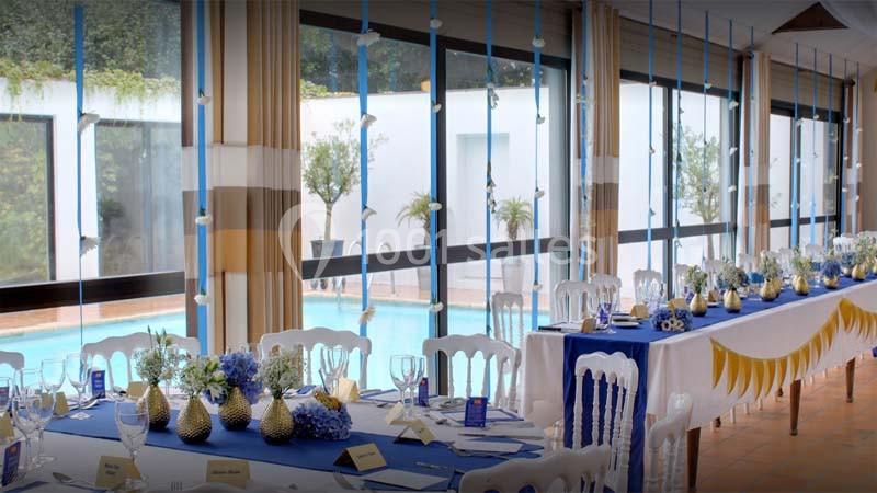Salle de réception lumineuse avec tables décorées de nappes bleues, centres de table fleuris et vue sur une piscine…
