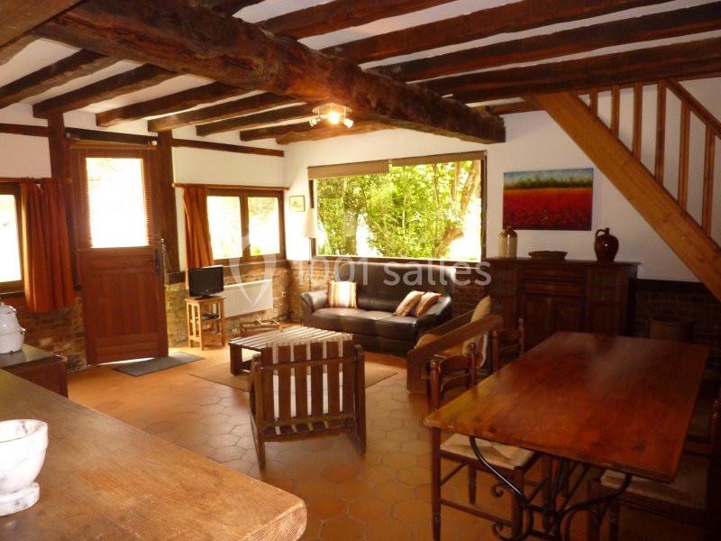 Salon rustique avec poutres apparentes, mobilier en bois, canapé, table à manger et vue sur un jardin verdoyant.
