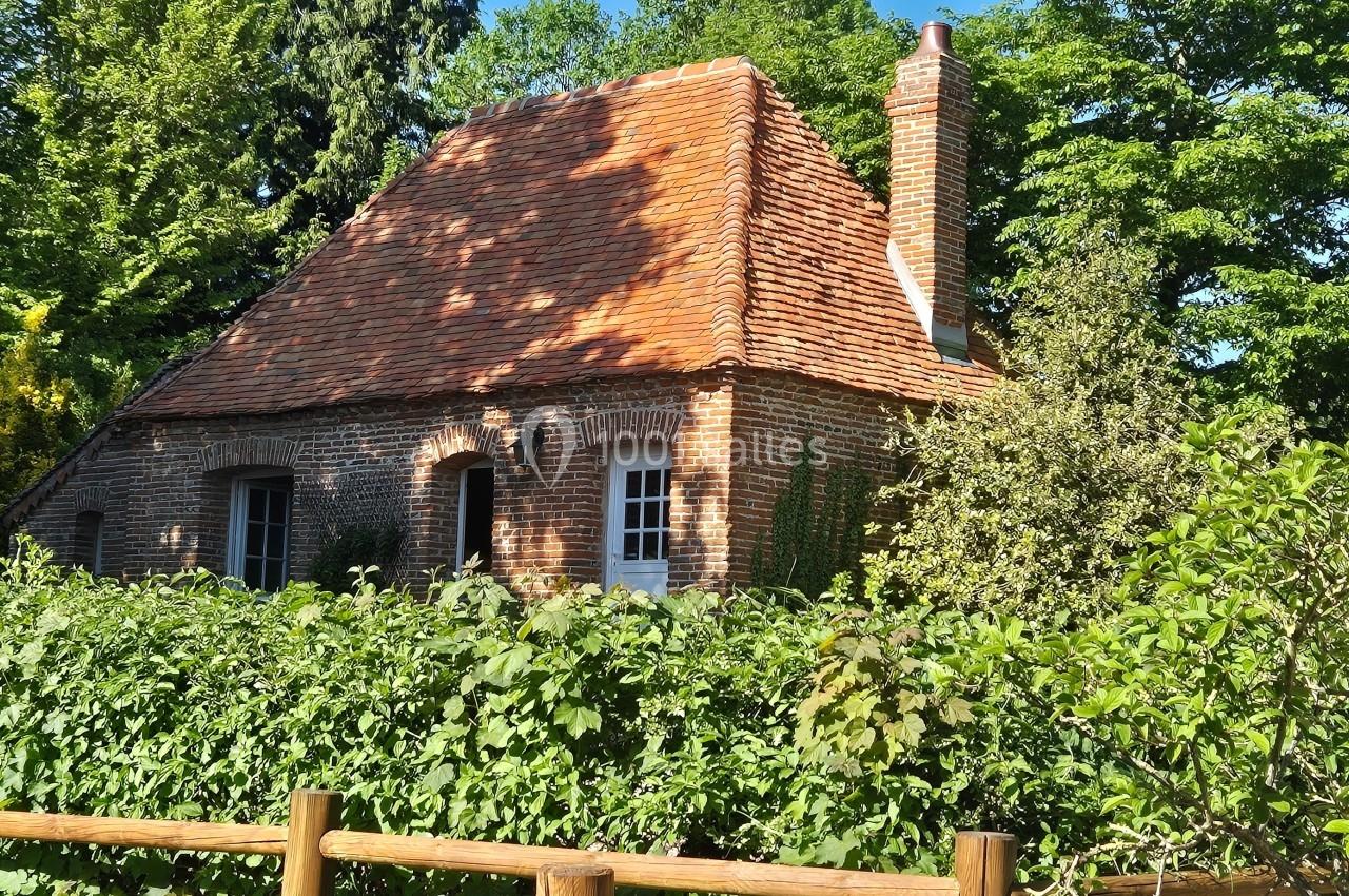 Maison en briques rouges avec toit en tuiles, entourée de verdure et d'une haie, par une journée ensoleillée.