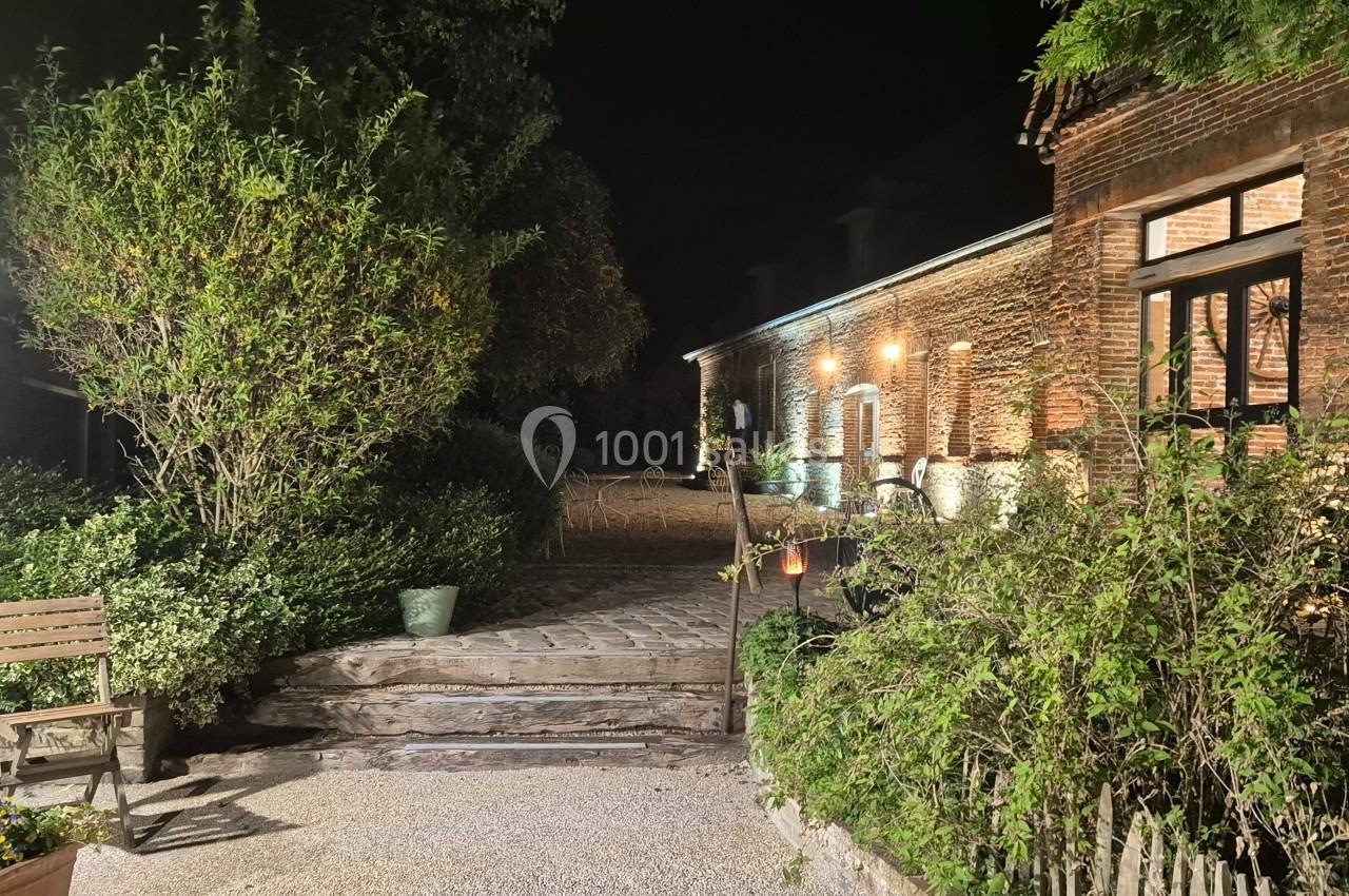 Cour extérieure éclairée de nuit, avec un bâtiment en briques, des plantes et un banc en bois.