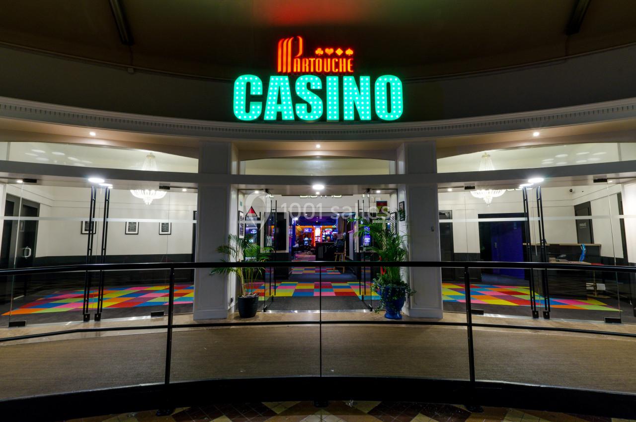 Entrée éclairée d'un casino avec enseigne lumineuse, sol coloré et portes vitrées donnant sur l'intérieur.