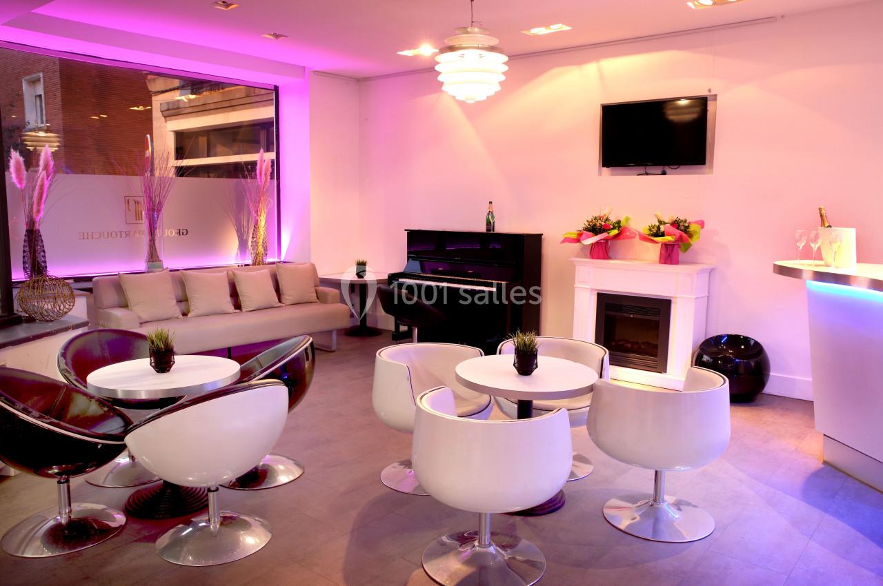 Salon moderne avec canapé, piano, tables rondes, chaises blanches et éclairage rose, décoré de plantes et fleurs.